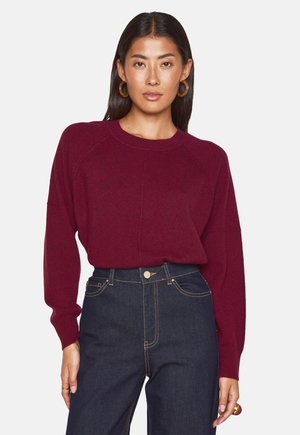 Bordeaux gebreide trui met geribbelde ronde hals en lange raglanmouwen, gecombineerd met donkerdenimjeans met hoge taille en knoopsluiting.