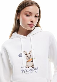 Witte hoodie met een geborduurde konijn die een wortel vasthoudt, met de tekst "Altijd blij zijn" en een ronde patch "eet vers".