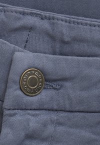 Blauer Stoff mit feiner Textur und sichtbaren Nähten, ausgestattet mit einem runden Metallknopf mit eingraviertem Markennamen.