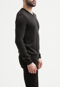 Czarny sweter w serek wykonany z miękkiego materiału, z długimi rękawami i ribbowanymi mankietami, zaprojektowany w celu uzyskania dopasowanego kroju. Widoczne minimalne detale zszycia.