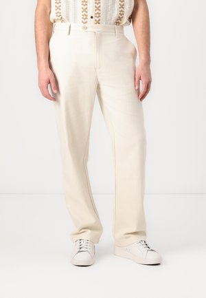 Pantaloni beige realizzati in tessuto morbido con taglio dritto, dotati di tasche frontali e passanti per cintura, abbinati a sneakers bianche.