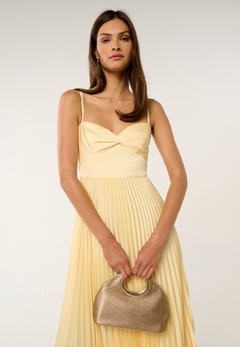 Vestido amarillo con pliegues, un escote anudado y tirantes finos; la modelo sostiene un bolso metálico dorado con un asa circular.