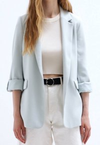 Žena v světle modrém blejzru přes bílý crop top, s vysokým pasem v bílých kalhotách a černým opaskem s kulatou sponou.