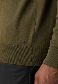 Maglione lavorato a maglia verde oliva con orlo e polsini a costine, caratterizzato da un piccolo tag in metallo vicino al fondo. Tessuto morbido, design aderente.