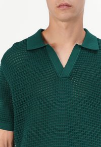 Polo en maille verte avec un col en V, des manches courtes et un motif texturé en tissage ouvert. Le col est uni et contrasté.