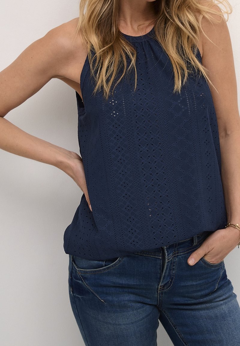 Kvinna i ärmlös marinblå strukturerad topp och blå jeans, med ena handen på höften och den andra i fickan, stående mot en enkel vägg.