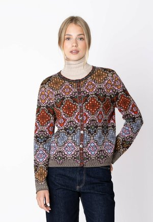 Young woman wearing a colorful patterned buttoned cardigan over a beige turtleneck, paired with dark blue jeans, standing against a white background.