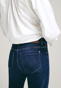 Jeans in denim blu scuro con una vestibilità su misura, dotati di tasche posteriori e dettagli di cucitura discreti, indossati con una camicia bianca a maniche lunghe.