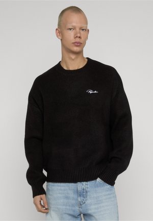 Nuori mies, jolla on lyhyet vaaleat hiukset, musta neulepaita, jossa on valkoista tekstiä rinnassa, ja vaaleansiniset farkut, yksinkertaisen taustan edessä.