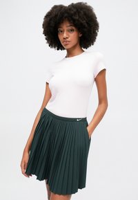 Jasnoróżowa koszulka zestawiona z ciemnozieloną spódnicą z zakładkami. Spódnica ma elastyczny pas i małe widoczne logo z przodu.