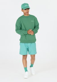 Grön sweatshirt med ribbade ärmar, tillsammans med ljusblå shorts. Vita sneakers och grön keps fulländar looken. Enkla logotyper på tillbehören.