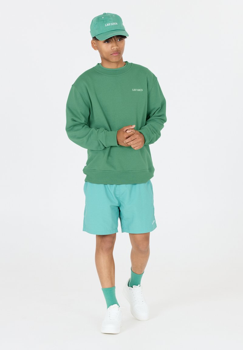 Grön sweatshirt med ribbade ärmar, tillsammans med ljusblå shorts. Vita sneakers och grön keps fulländar looken. Enkla logotyper på tillbehören.