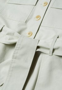 Gros plan d'une chemise beige avec des boutons sur le devant, deux poches poitrine et une large ceinture en tissu assorti nouée à la taille.