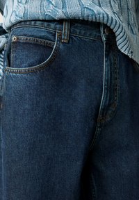 Denim kavbojke z temno modrim pranjem. Značilnosti vključujejo kontrastno oranžno šivanje, klasičen model s petimi žepi in gladko teksturo.