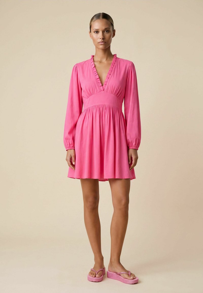 Femme aux cheveux plaqués en arrière portant une robe rose à manches longues, longueur genoux, et des tongs compensées roses, debout devant un fond beige.