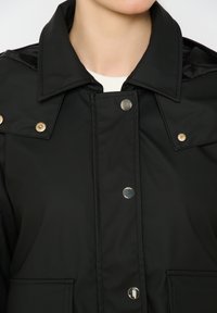 Juodas paminkštintas švarkas su dideliu apykakle, sidabriniais mygtukais ir užtrauktuku. Jis turi lygią tekstūrą ir šonines kišenes.