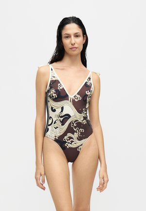 Femme aux cheveux mouillés porte un maillot de bain une pièce marron et crème avec un motif abstrait ondulé et des fines bretelles sur un fond uni.