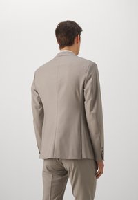 Veste de costume taillée gris clair avec revers à cran et une fente arrière. Dotée de quatre boutons sur les poignets et d'une texture lisse et structurée.