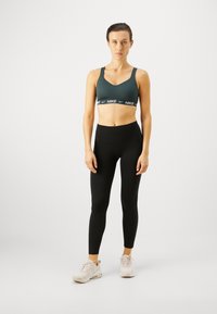 Sujeta oscura de deporte verde con banda negra con el logo de Nike, combinada con leggings negros de cintura alta y zapatillas deportivas rosas claras.