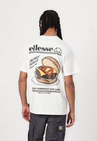 Camiseta blanca con un gráfico de una hamburguesa con una rebanada de queso amarillo y salsa roja. La parte posterior muestra el logo de "ellesse" y texto.