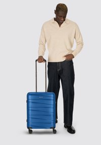 Uomo in maglione beige e jeans scuri che tiene il manico di una valigia blu con rotelle, guardando in basso, in piedi su uno sfondo grigio chiaro.