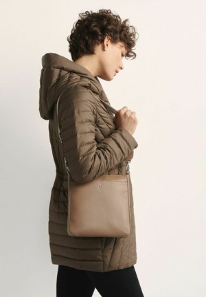 Kobieta z krótkimi kręconymi włosami, ubrana w brązowy pikowany płaszcz i niosąca beżową skórzaną torbę na ramieniu, na jednolitym tle.