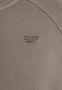 Cálido pulóver de Reebok en un suave tono marrón apagado. Presenta una textura acanalada con un escote redondo y un logo bordado en el pecho.