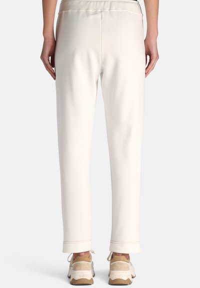 Personne portant un pantalon droit couleur crème et des baskets beiges, vue de dos sur un fond uni.