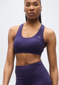 Γυναίκα με μοβ αθλητικό σουτιέν και κολάν, στέκεται μπροστά σε απλό φόντο, δείχνοντας τον τονωμένο κορμό και τα χέρια της.