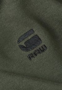 Olijfgroene stof met een geborduurd zwart logo, dat een gladde textuur en een eenvoudig geometrisch ontwerp toont.