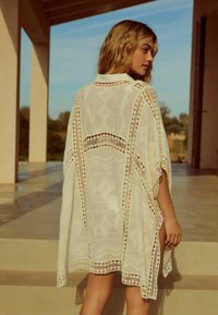 Kimono blanc léger avec des détails en dentelle, manches amples et une texture à motifs, positionné en extérieur sur un fond neutre.