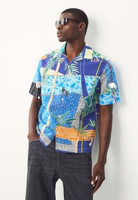 Chemise à manches courtes, boutonnée, de couleur bleue avec des motifs tropicaux éclatants, comprenant des palmiers et des dessins géométriques. Portée avec un pantalon en denim foncé.