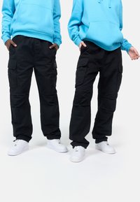 Black cargo pants with multiple pockets, paired with a light blue hoodie. Both wear white sneakers. Casual and relaxed fit design.