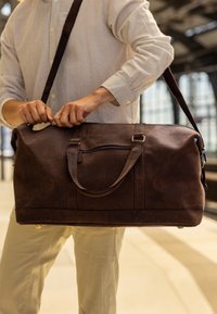 Brown leather duffel bag with dual handles and removable shoulder strap, featuring a zippered top and front pocket for storage.