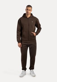 Sudadera con cremallera marrón, con bolsillos frontales y parche de logo, combinada con pantalones de chándal a juego. Zapatillas blancas completan el conjunto. Textura suave y lisa.