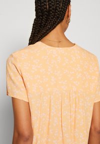 Mujer con cabello trenzado que lleva una blusa de manga corta color durazno con un pequeño estampado floral blanco, vista desde atrás.