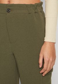 Pantaloni affusolati verde oliva con vita arricciata, chiusura con bottone, tasche laterali e texture del tessuto liscia.