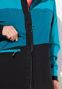 Veste teal et noire avec des poches zippées, un col montant et un tissu texturé. Présente des blocs de couleurs contrastées et un détail de marque.