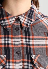 Camicia in flanella con motivo a quadri nero, bianco e arancione, design con bottoni e colletto, con taschini sul petto a contrasto. Texture morbida.