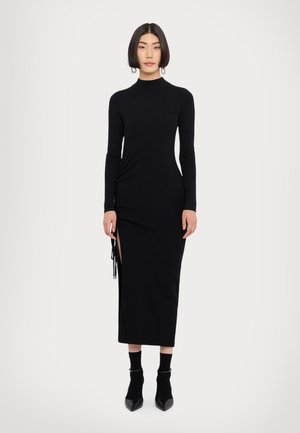 Black ribbed knit dress with a high neck, long sleeves, side slit, and adjustable fringe detailing. Paired with ankle-length black socks and heels.