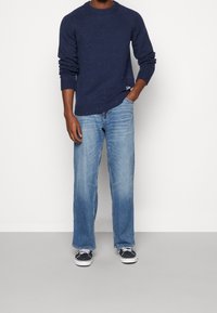 Man med en marinblå spräcklig tröja, ljusblå raka jeans, svart och vit sneakers, står med ena handen i fickan.