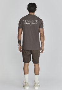 Brūna t-kreklā ar baltu tekstu, kas rāda "SIKSILK Follow the Movement", apvienota ar saskaņotām šortiem un baltām zeķēm, stāv uz vienkārša fona.