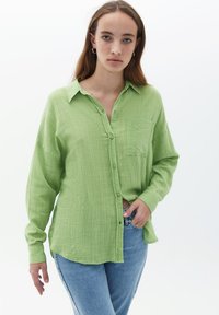 Zielona koszula z fakturowanego materiału, z kołnierzykiem i kieszonką na piersi, w połączeniu z niebieskimi dżinsami. Miękki, lekki materiał o luźnym kroju.