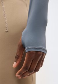 Κοντινό πλάνο ενός σκούρου δέρματος χεριού που φοράει μπλε ρούχο με μακριά μανίκια και τρύπα για τον αντίχειρα, δίπλα σε μπεζ παντελόνι με ορατή ραφή.