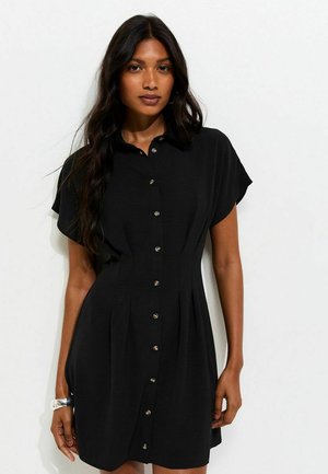 Femme aux longs cheveux foncés portant une robe noire à manches courtes avec des boutons et des plis, debout devant un fond clair uni.