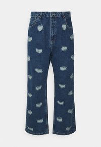 Jeans de denim em azul escuro com um corte largo, cintura alta e vários remendos desgastados com bordas esfiapadas.