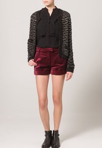 Schwarze Strickjacke mit metallischen Akzenten, Bluse mit Rüschendetails, burgunderrote Samtshorts und schwarze Ankle Boots.