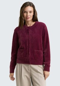 Bordeaux, fluffy cardigan met een ronde halslijn, voor knoopsluiting en twee opgestikte zakken op de taille. Zachte textuur, lange mouwen.