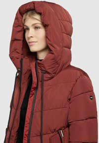 Giacca imbottita rossa con un grande cappuccio trapuntato, cerniera nera e dettagli con coulisse, caratterizzata da una texture elegante e tasche laterali con zip.