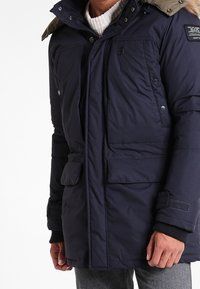 Mörkblå vadderad jacka med avtagbar huva, utrustad med två fickor fram, tryckknappar och en strukturerad stickad krage.